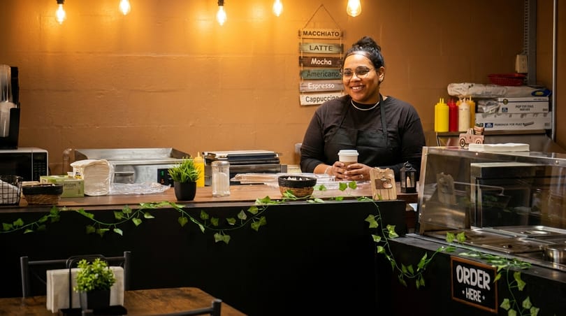 Smiling barista in an apron holding a coffee cup behind a counter with a menu board.